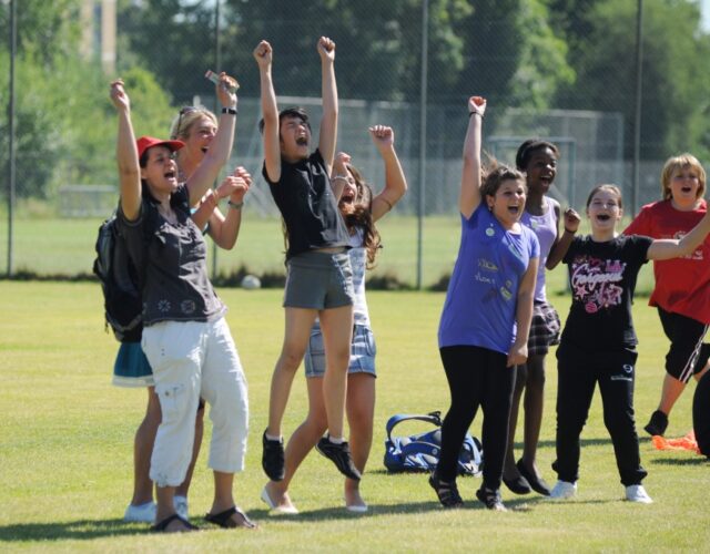 Jugendliche stehen jubelnd auf eine Sportplatz. Sie rufen freudig und strecken die Hände in die Luft.